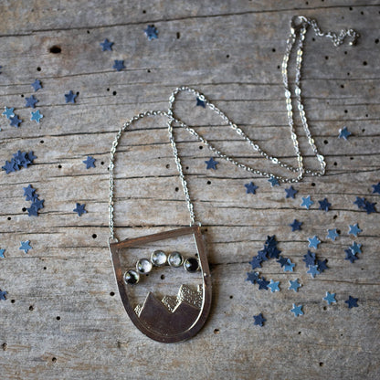 A silver shield like pendant with a mountain scene and 5 moon phases overhead, resting on a wooden background with blue star confetti. 