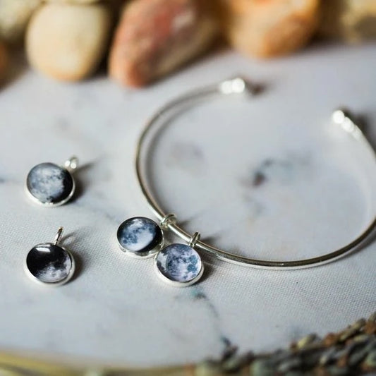 A silver-toned open cuff bracelet with circle moon phase charms on a light marble surface with blurred stones in the background.