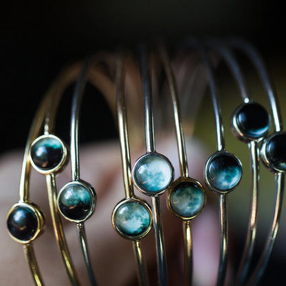 A close-up of several thin gold-toned bangle bracelets with moon phase cabochons, shown against a dark blurred background.