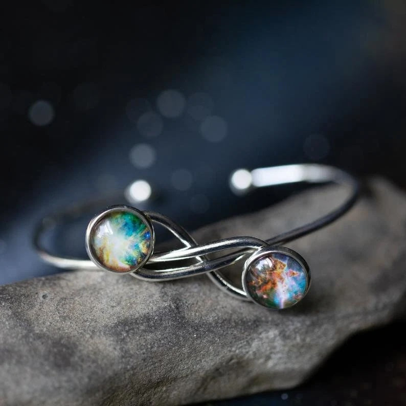 A silver infinity-style cuff ring with two round galaxy cabochons, resting on a textured gray stone against a dark, blurred background.