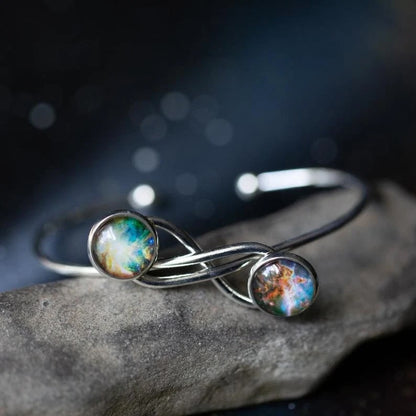 A silver infinity-style cuff ring with two round galaxy cabochons, resting on a textured gray stone against a dark, blurred background.