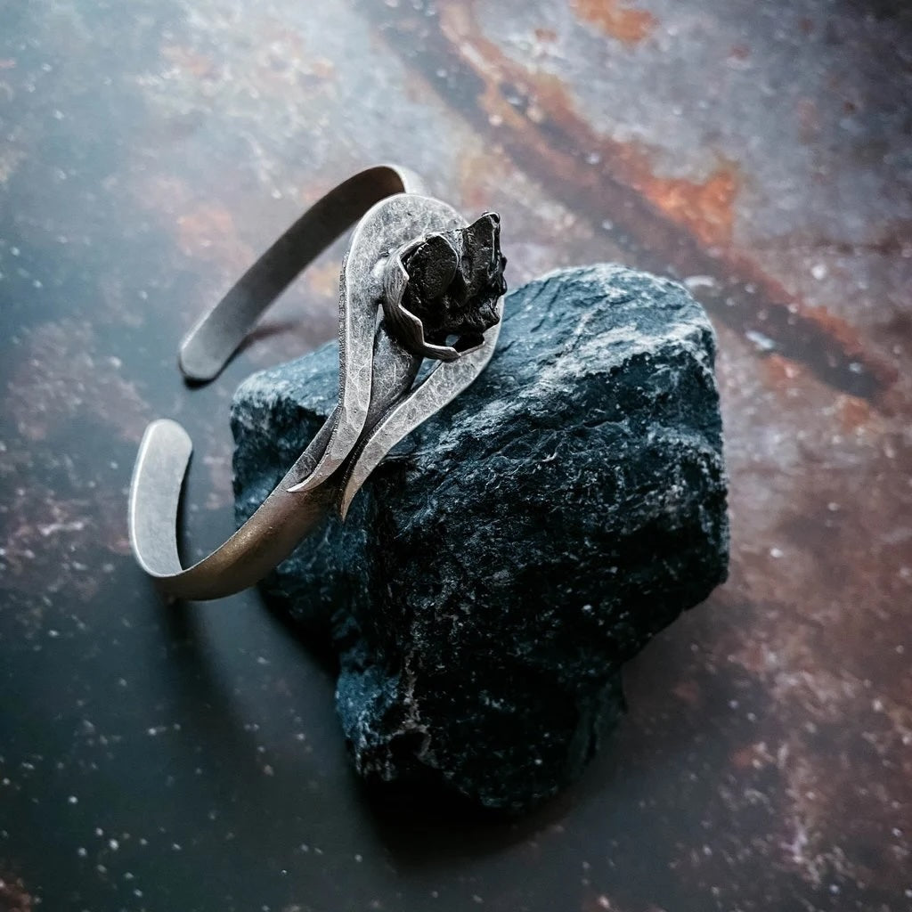 A handmade silver-toned metal cuff bracelet set with authentic meteorite, displayed on a dark, rough stone against a dark, rustic background.