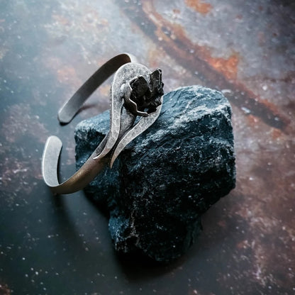 A handmade silver-toned metal cuff bracelet set with authentic meteorite, displayed on a dark, rough stone against a dark, rustic background.