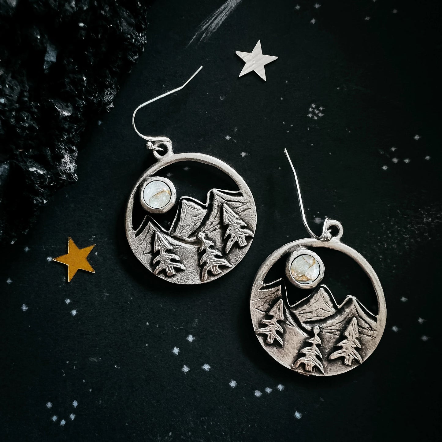 A pair of silver-toned round drop earrings featuring an engraved mountain and pine tree design with a small oval stone, displayed on a dark starry background.