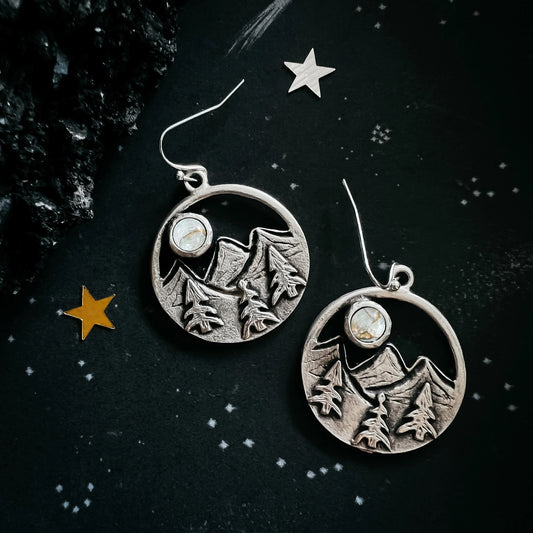 A pair of silver-toned round drop earrings featuring an engraved mountain and pine tree design with a small oval stone, displayed on a dark starry background.