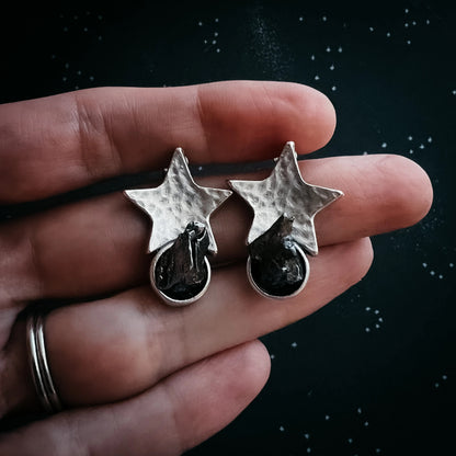 A pair of silver-toned star-shaped earrings with dark meteorite set below, held on a person's hand against a dark starry background.