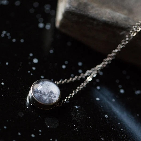 A silver-toned chain necklace with a round pendant featuring a moon design inside a clear dome, displayed on a dark, starry surface next to a stone.