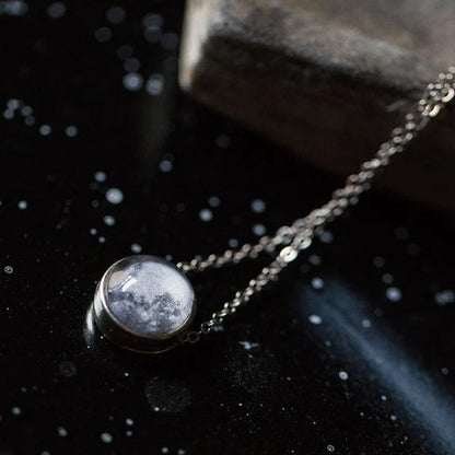 A silver-toned chain necklace with a round pendant featuring a moon design inside a clear dome, displayed on a dark, starry surface next to a stone.