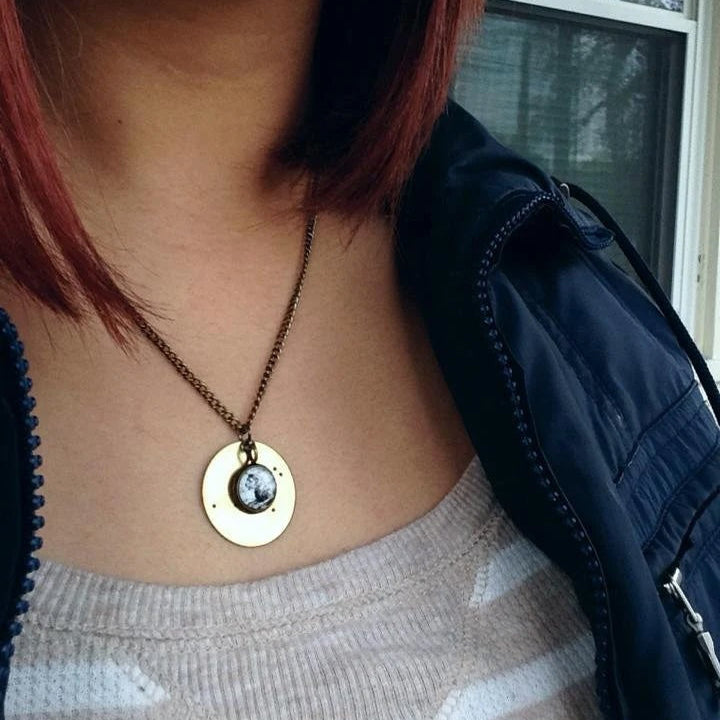 A close-up of a person wearing a circular pendant necklace with a gold-toned disc and a small circle cabochon, paired with a dark navy jacket and a light gray top.