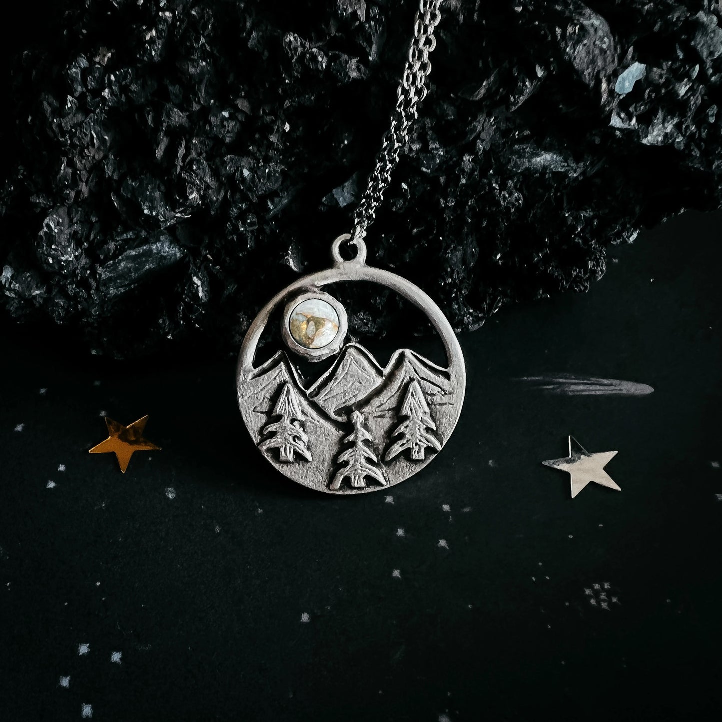 A silver-toned circular pendant necklace featuring an engraved mountain and pine tree design, with a small round stone inset near the top, hanging from a chain against a dark textured background with decorative star-like props.