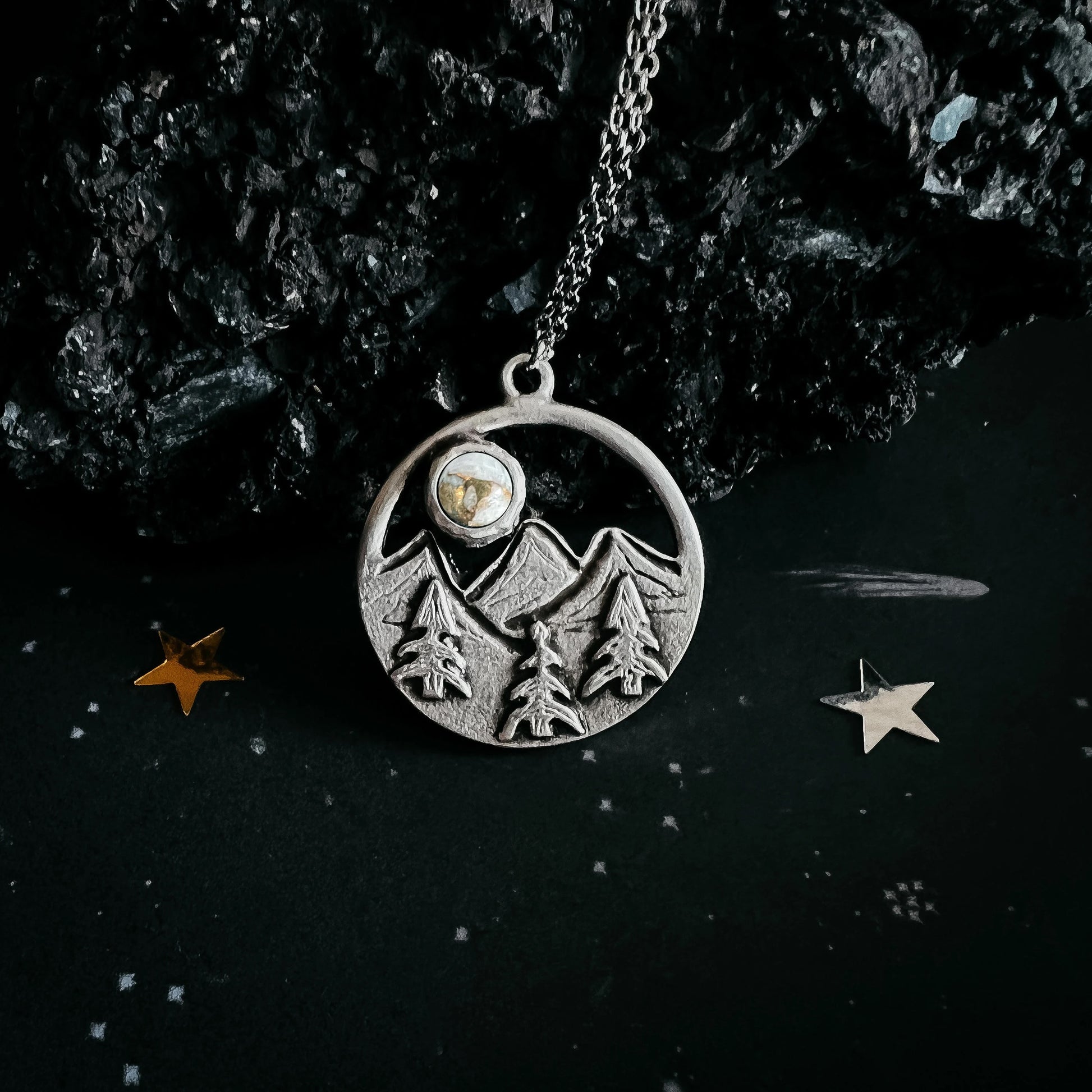 A silver-toned circular pendant necklace featuring an engraved mountain and pine tree design, with a small round stone inset near the top, hanging from a chain against a dark textured background with decorative star-like props.