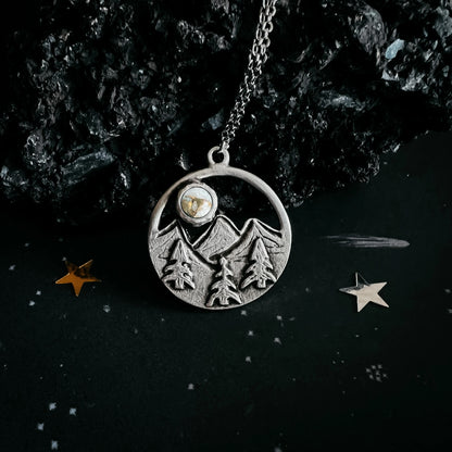 A silver-toned circular pendant necklace featuring an engraved mountain and pine tree design, with a small round stone inset near the top, hanging from a chain against a dark textured background with decorative star-like props.