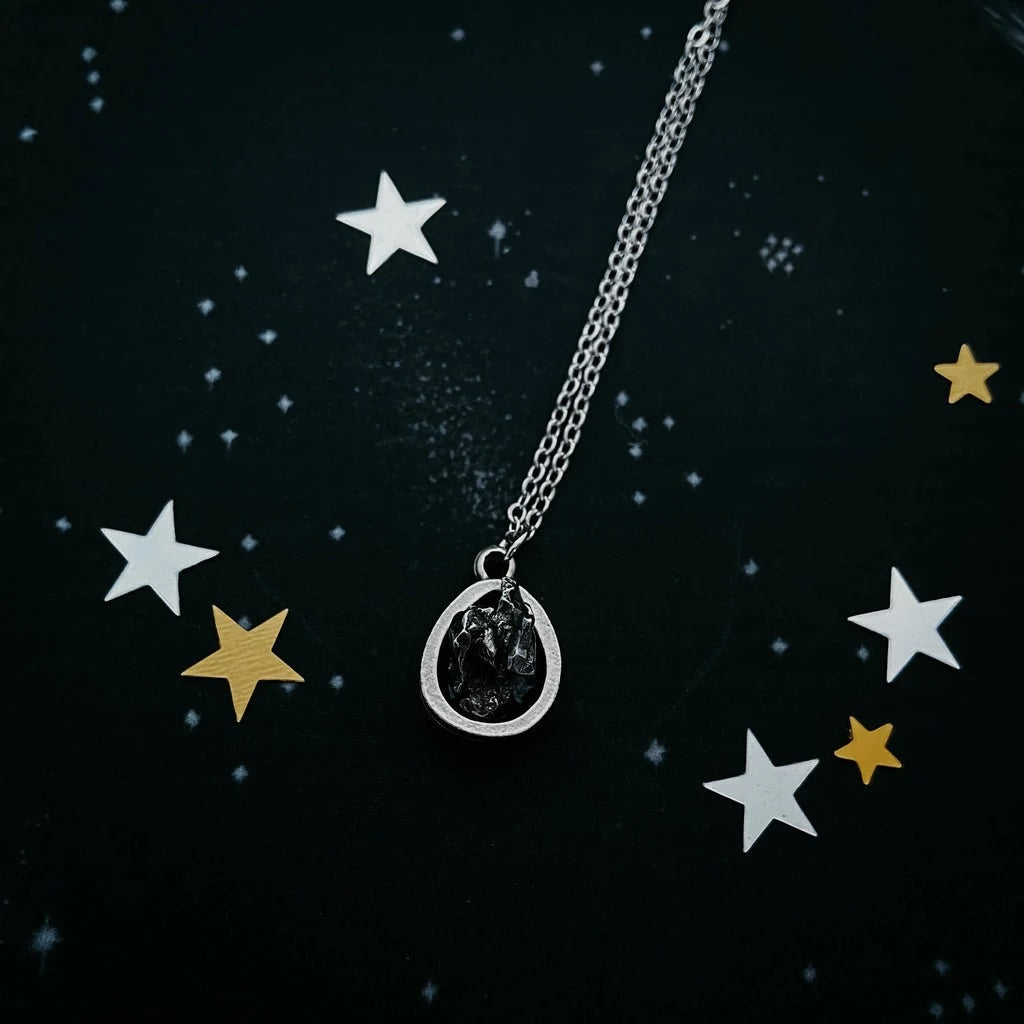 A silver chain necklace with a teardrop-shaped pendant featuring a dark, textured central design surrounded by a silver border, displayed on a dark starry background with white and gold star confetti.