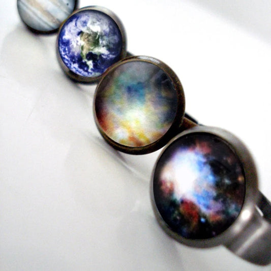 A close-up of a silver and bronze toned rings featuring different galaxy and planet themed cabochons, displayed on a white surface.