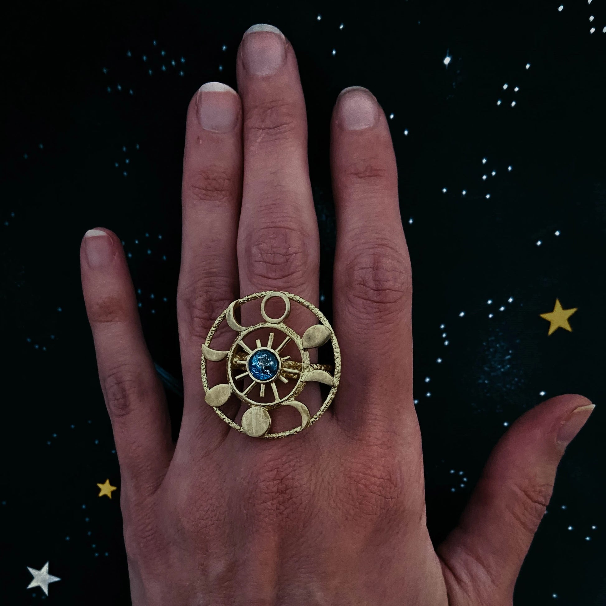 Hand wearing a gold ring with an earth image at the center against a starry night sky background