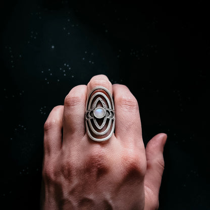 A hand wearing a large oval silver ring with concentric geometric bands and a central white stone, set against a dark starry background.