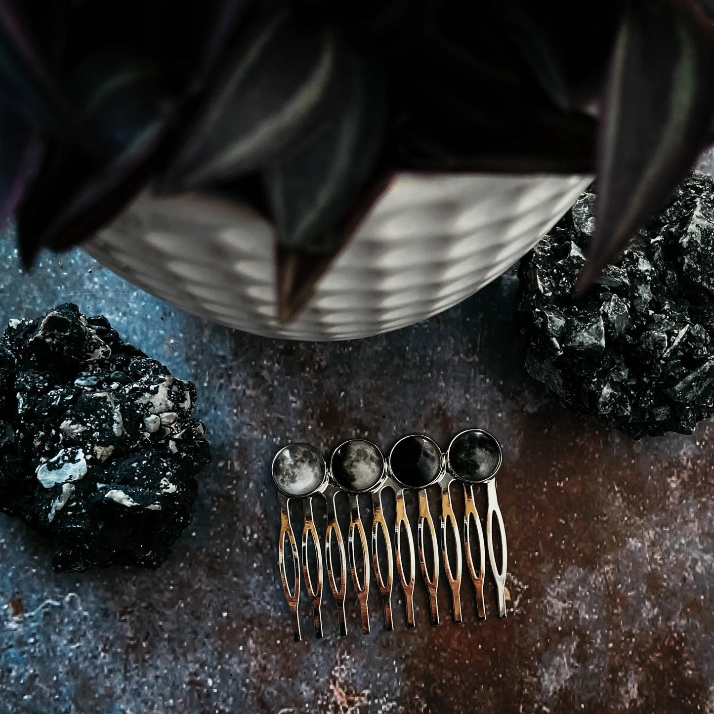 A four moon phase comb on a textured dark surface with decorative rocks and a potted succulent in the background.