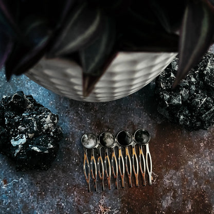 A four moon phase comb on a textured dark surface with decorative rocks and a potted succulent in the background.