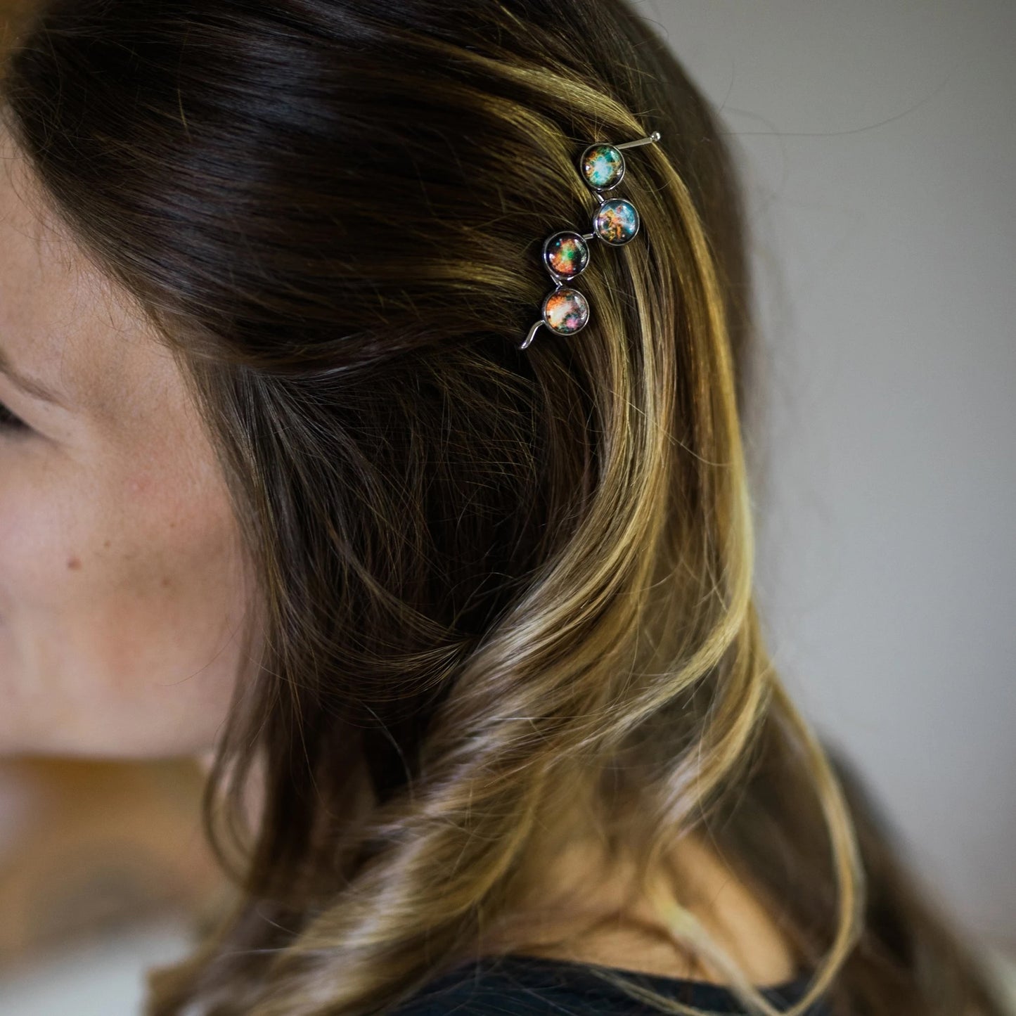 A close-up side profile of a woman with long, highlighted brown hair wearing a silver-toned hair clip adorned with three iridescent, multicolored galaxy cabochons.