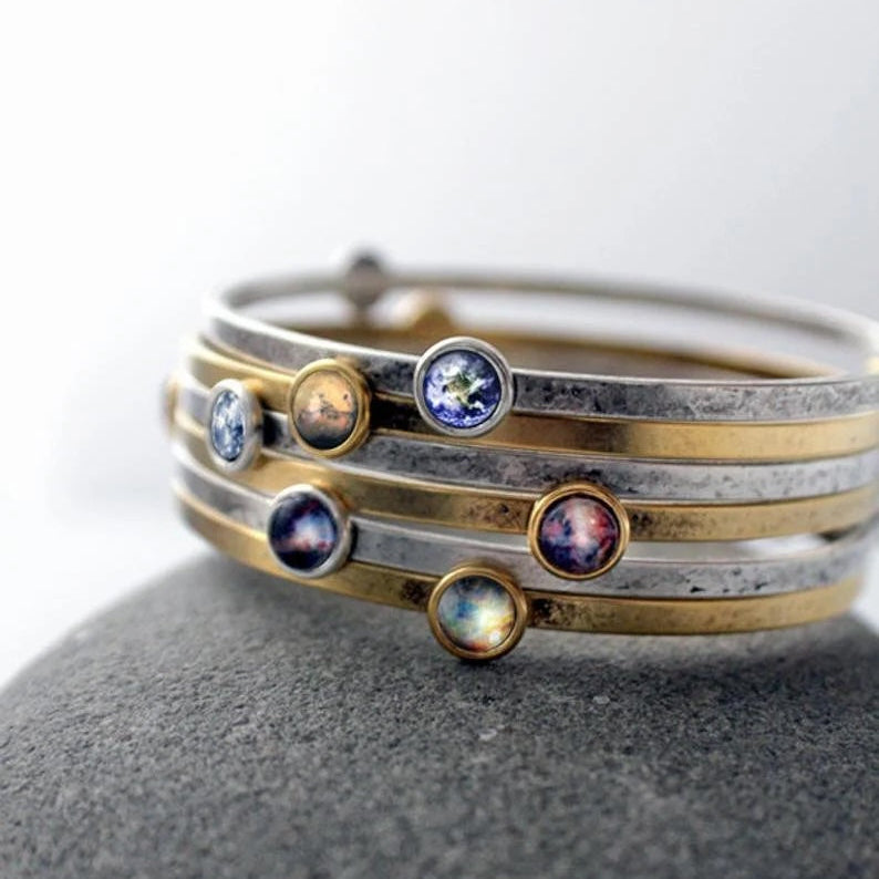 A stack of three thin metal bangles, including textured silver, polished gold, and mixed metal, each adorned with small round bezel-set gemstones with galaxy images, displayed on a dark gray stone against a white background.