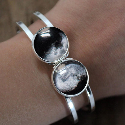A close-up of a person's wrist wearing a silver-toned cuff bracelet with two round cabochon stones showing a black-and-white moon phase design.