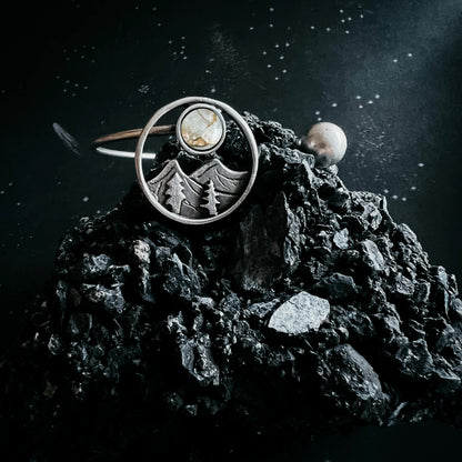 A silver-toned ring with a circular mountain and tree motif and a small oval stone, displayed on a dark textured rock against a starry black background.
