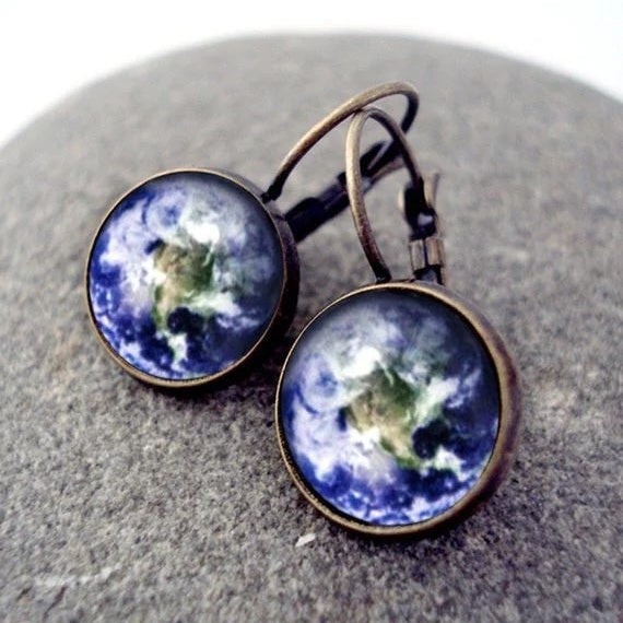 A pair of round drop earrings with a vintage bronze finish, featuring a detailed Earth design, displayed on a textured gray stone surface.