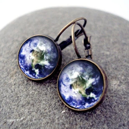 A pair of round drop earrings with a vintage bronze finish, featuring a detailed Earth design, displayed on a textured gray stone surface.