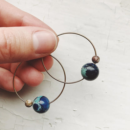 A close-up of a hand holding a pair of thin metal hoop earrings with round marbled beads in blue/green and white/brown coloring, against a light textured background.