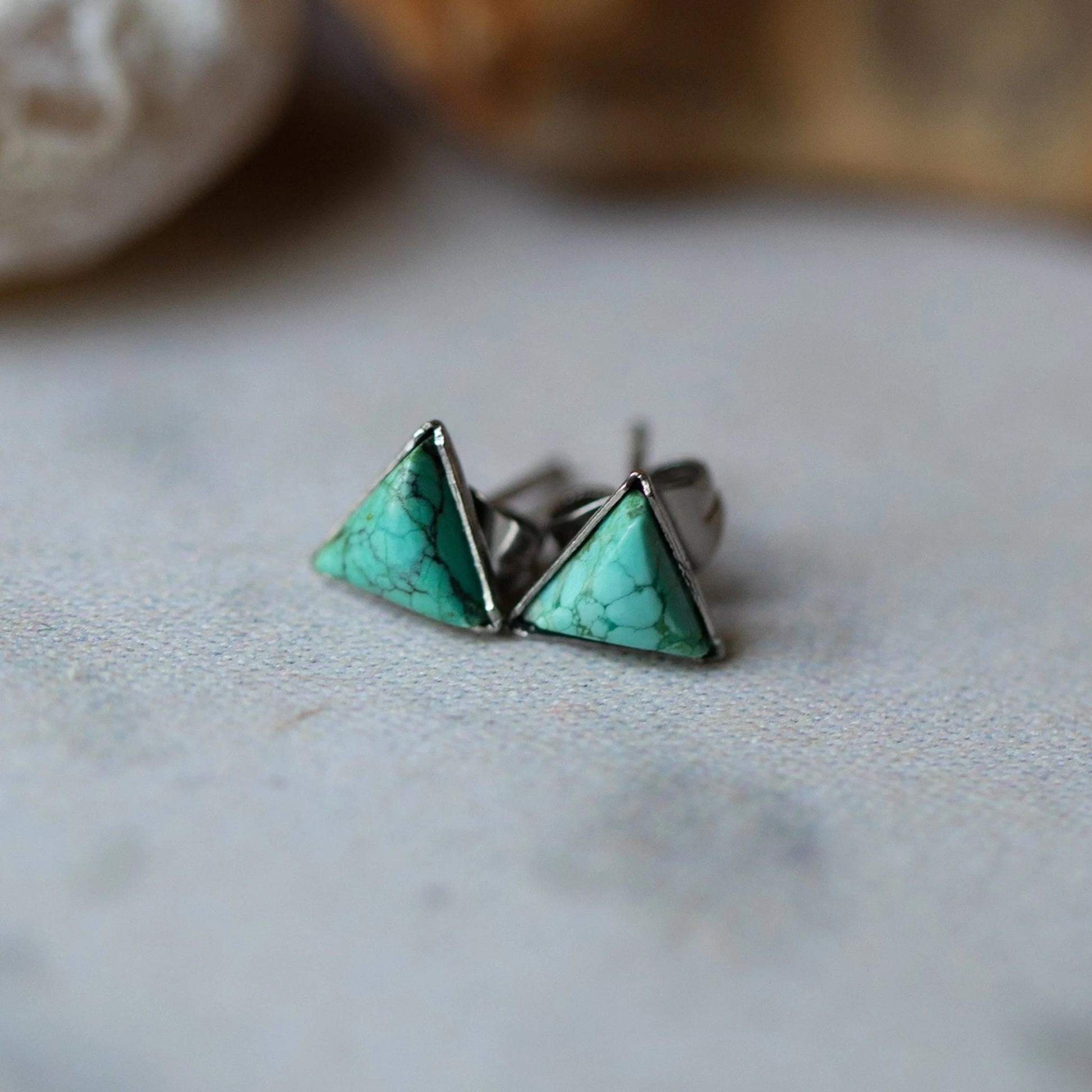 A pair of silver stud earrings with triangular turquoise stone inlays, resting on a textured light gray surface with a blurred natural background.