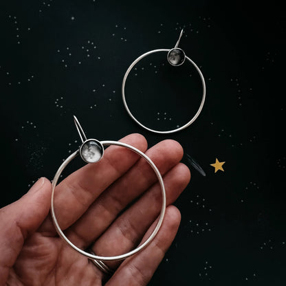 A pair of silver-toned hoop earrings with small circular moon cabochons, shown on a person's hand against a dark starry background.