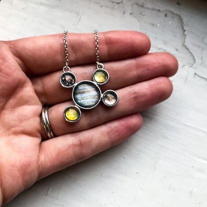 A necklace with a larger Jupiter image at the center center and smaller Jupiter's moons surrounding it, held in a person's hand. 