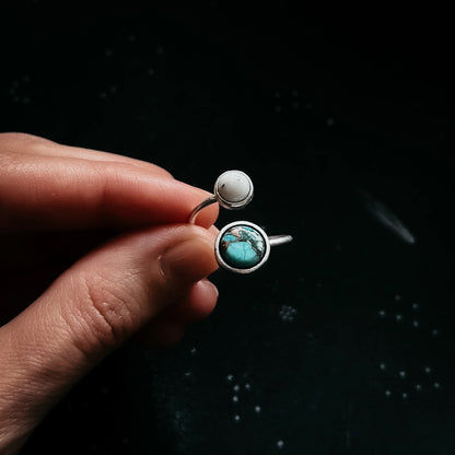 Hand holding a silver ring with a larger blue and smaller white stone that symbolize the earth and its moon, against a dark background