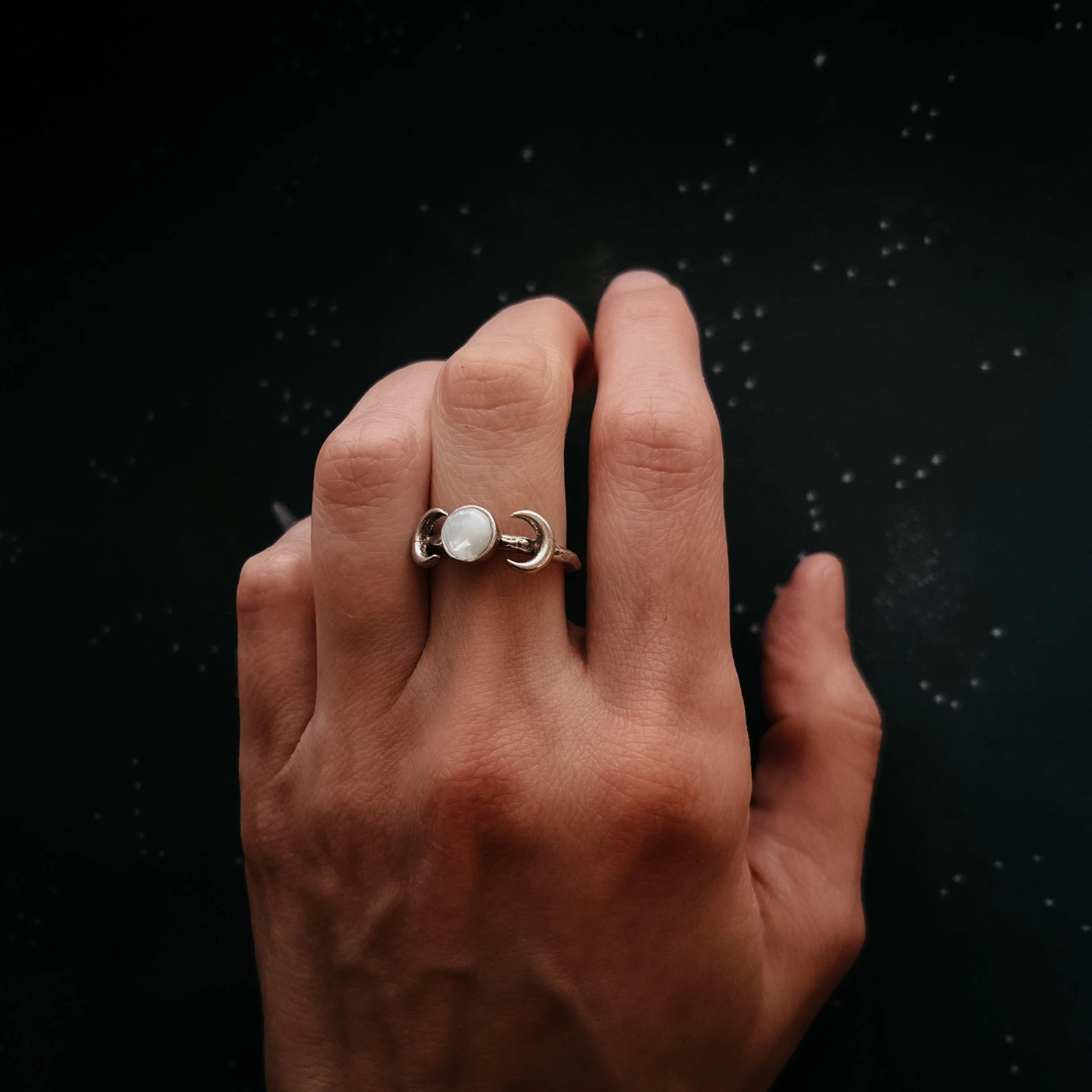 A silver ring with a central white moonstone, worn on a person's middle finger against a dark, speckled background.