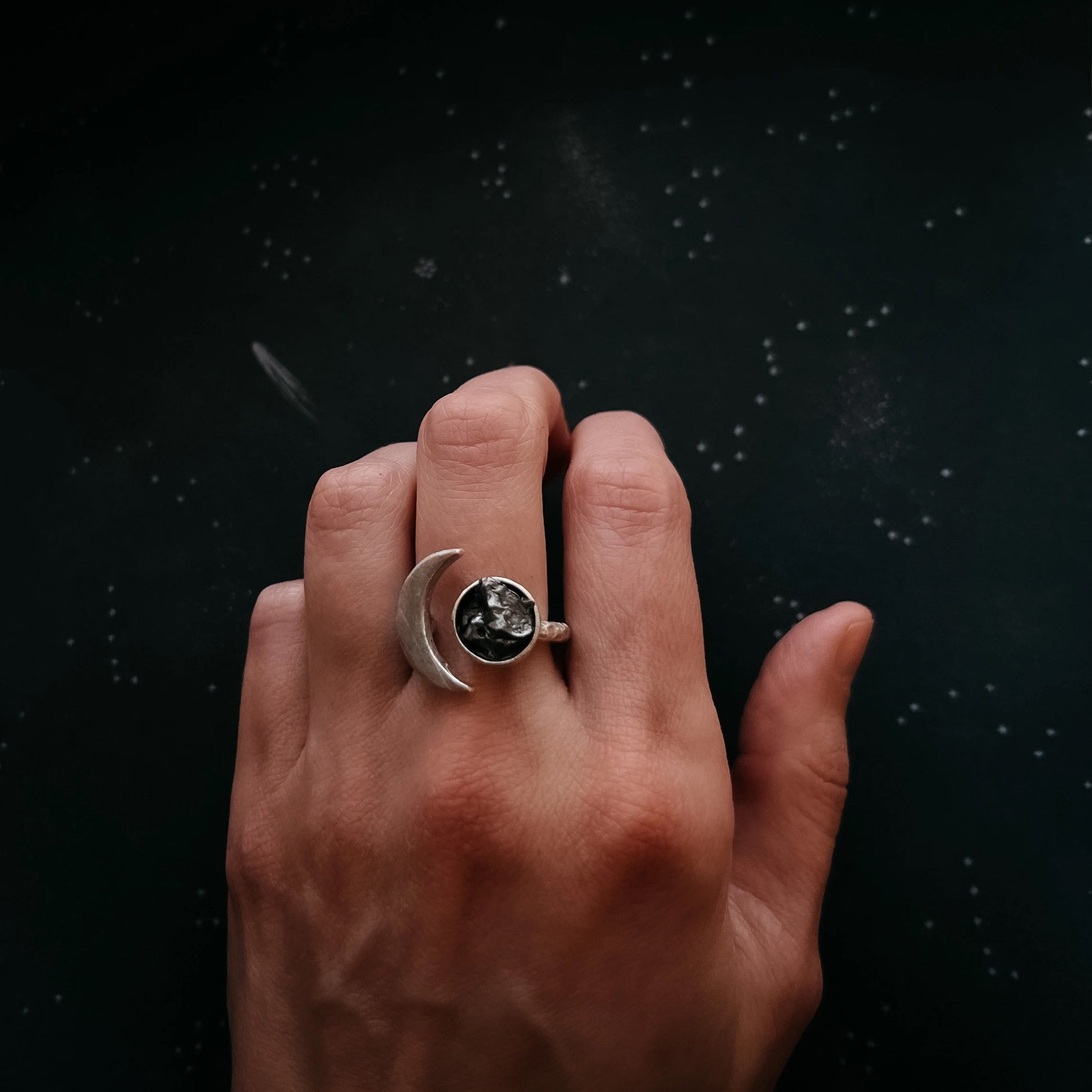 A hand wearing a silver crescent moon ring with a dark, textured meteorite stone, set against a dark, starry background.