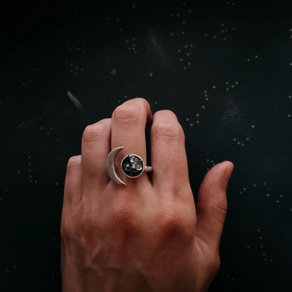 A hand wearing a silver crescent moon ring with a dark, textured meteorite stone, set against a dark, starry background.