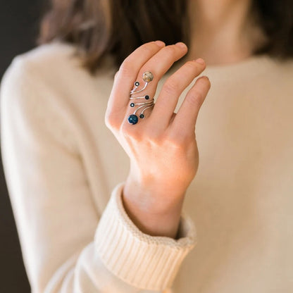 A close-up of a person's hand wearing a multi-stone silver ring with two larger blue and white stones and smaller black stones orbiting them, set against a soft cream-colored sweater.