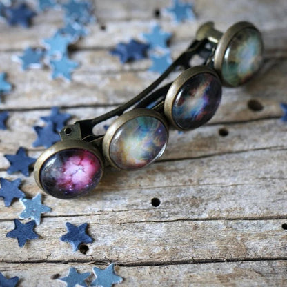 A small barrette with galaxy images set in its circles, resting on a rustic wooden surface scattered with blue star-shaped confetti.